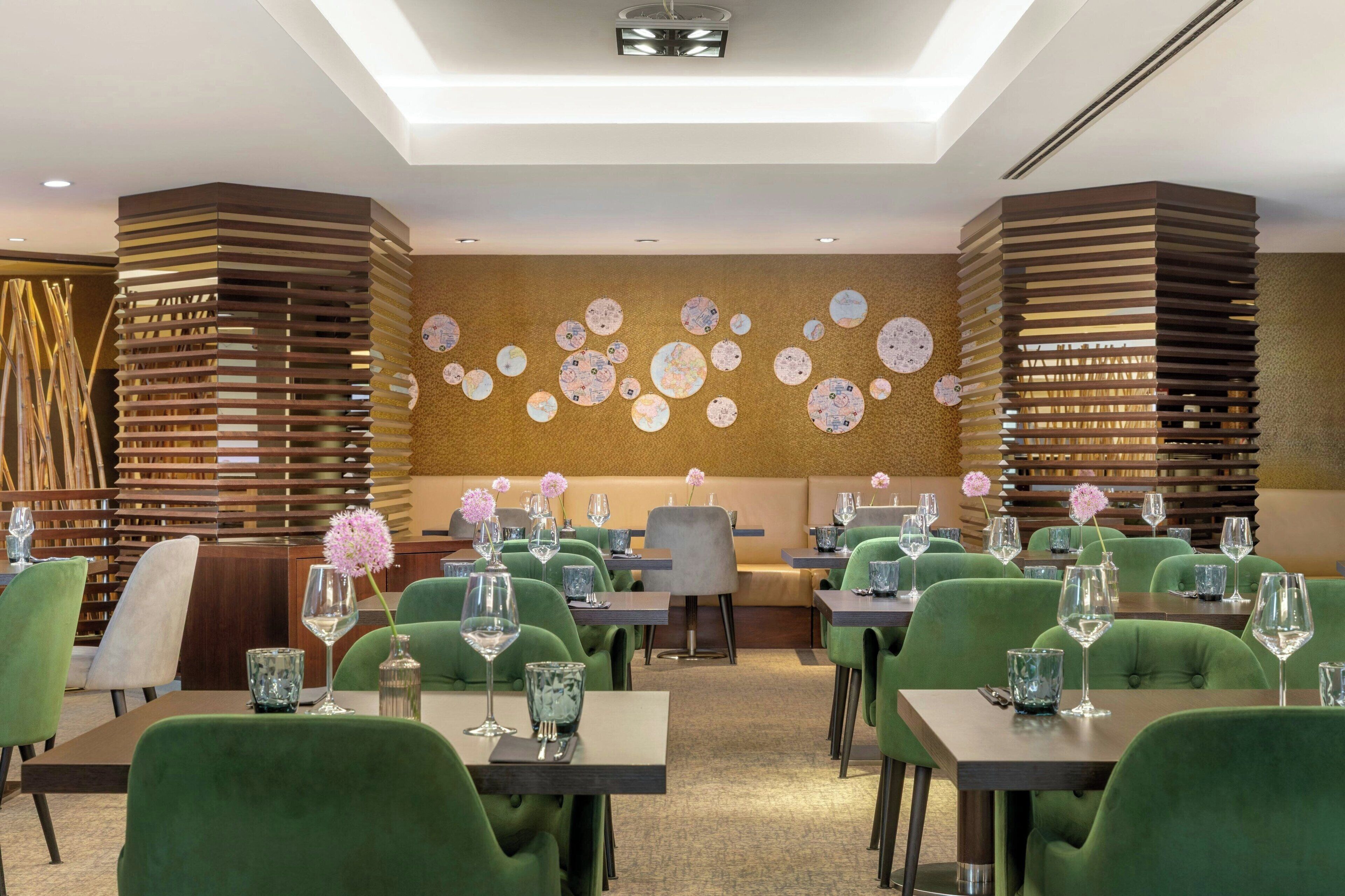 A modern restaurant with green upholstered chairs, tables with wine glasses, and decorative wall art.