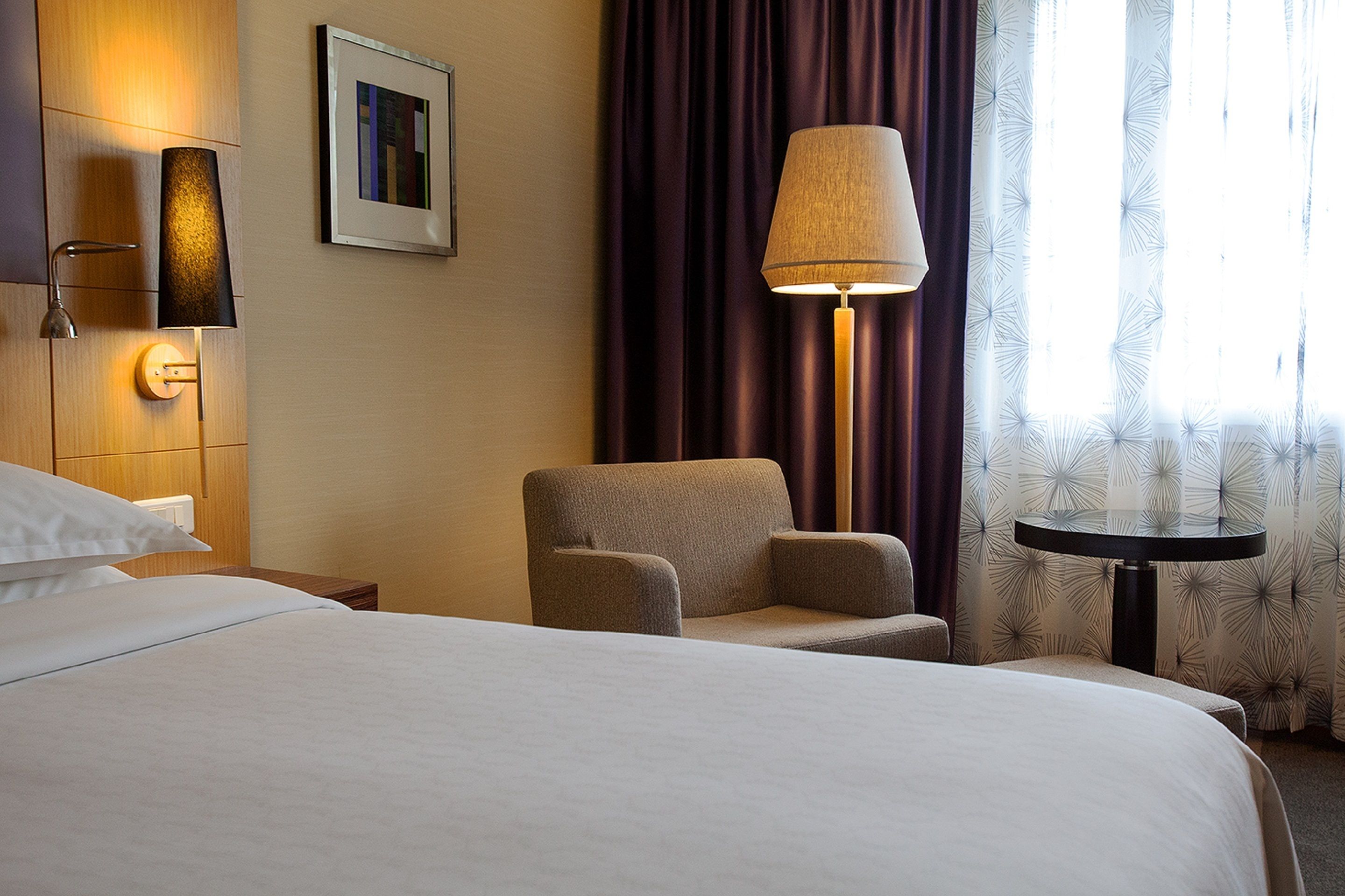 Une chambre d’hôtel comprenant un lit, une chaise, une table, une lampe, un luminaire fixé au mur et une fenêtre avec des rideaux.