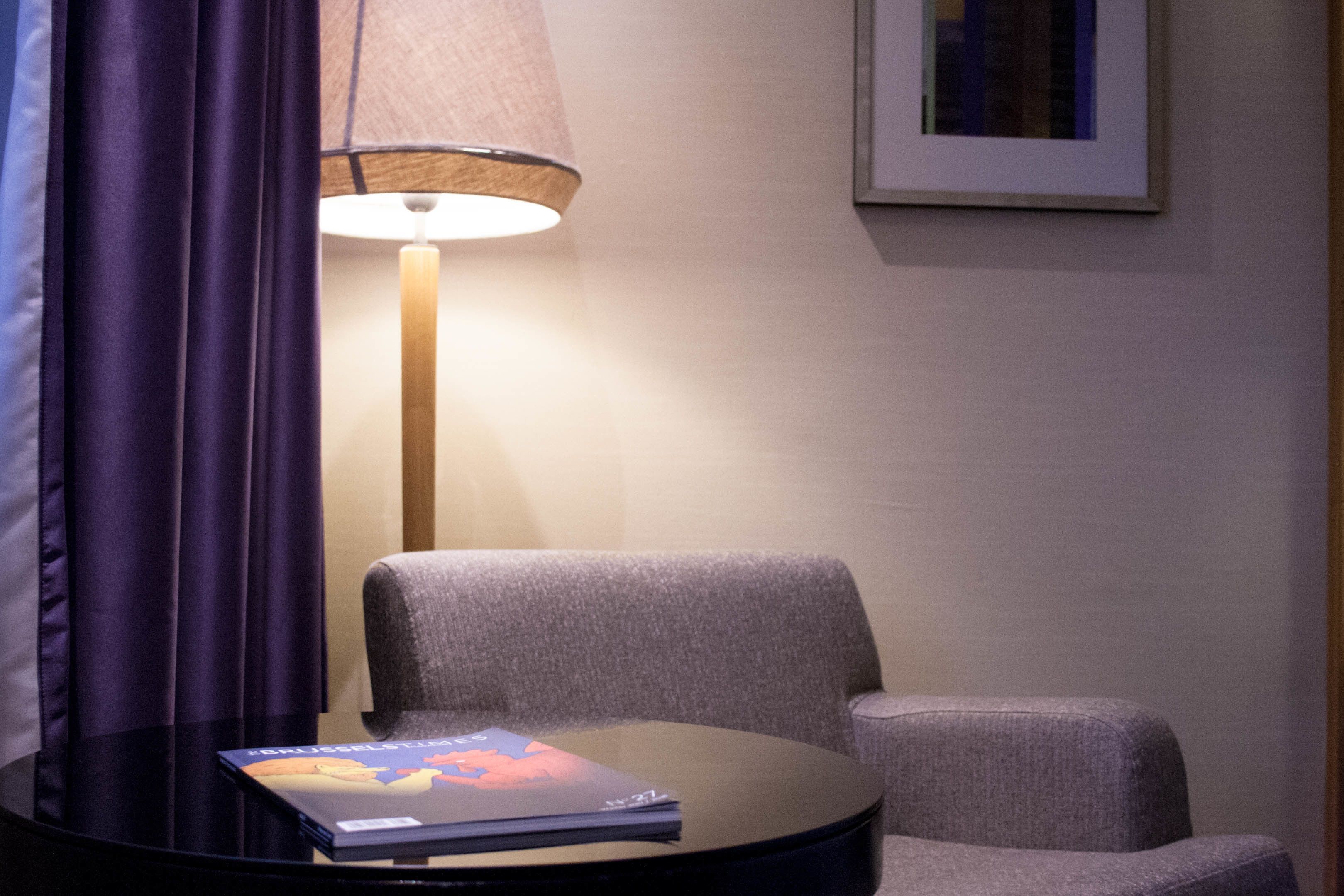 A room with a round table, a lamp, a chair, and a framed picture on the wall.