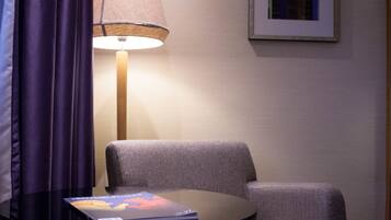 A room with a round table, a lamp, a chair, and a framed picture on the wall.