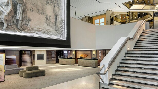 A spacious lobby with a high ceiling, a large mural, a staircase, and seating areas. - Sheraton Brussels Airport Hotel (Zaventem)