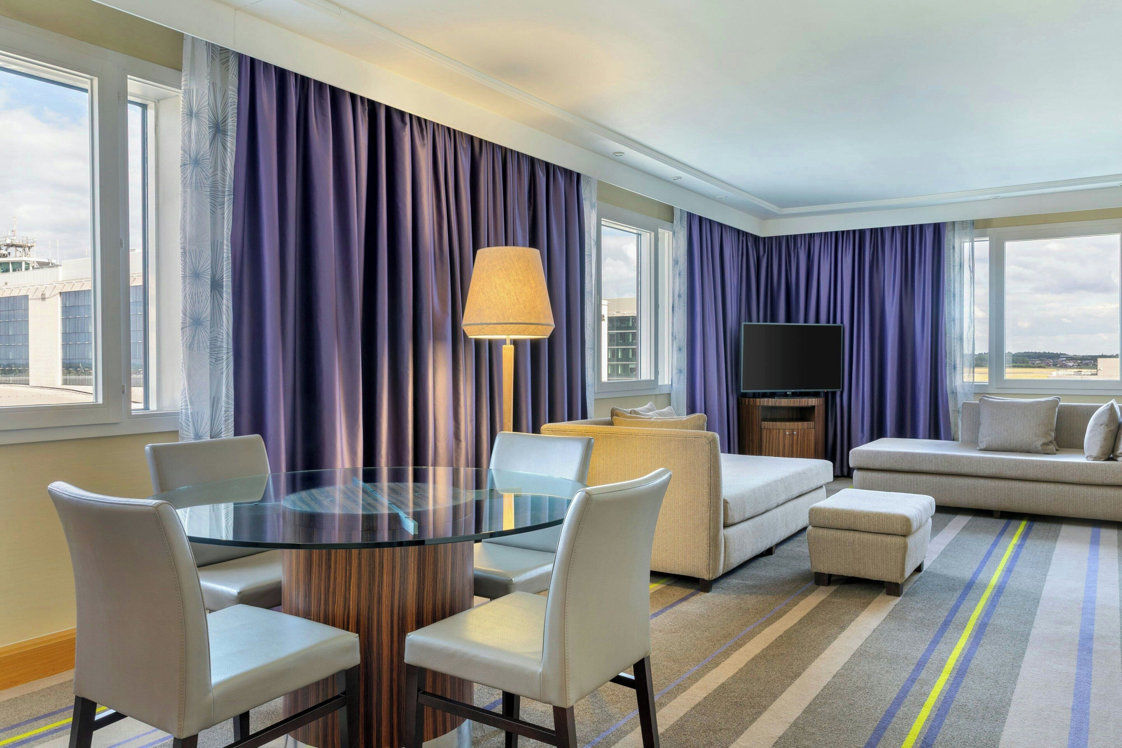 Habitación de hotel con una mesa de vidrio, dos sofás, un televisor y ventanas grandes con cortinas.