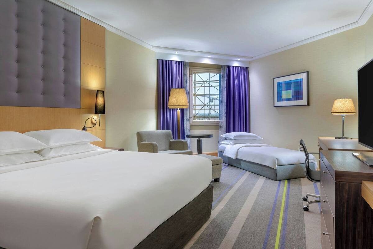 Une chambre d’hôtel avec un grand lit, un plus petit lit, un bureau, une chaise, une télévision et une fenêtre avec des rideaux.