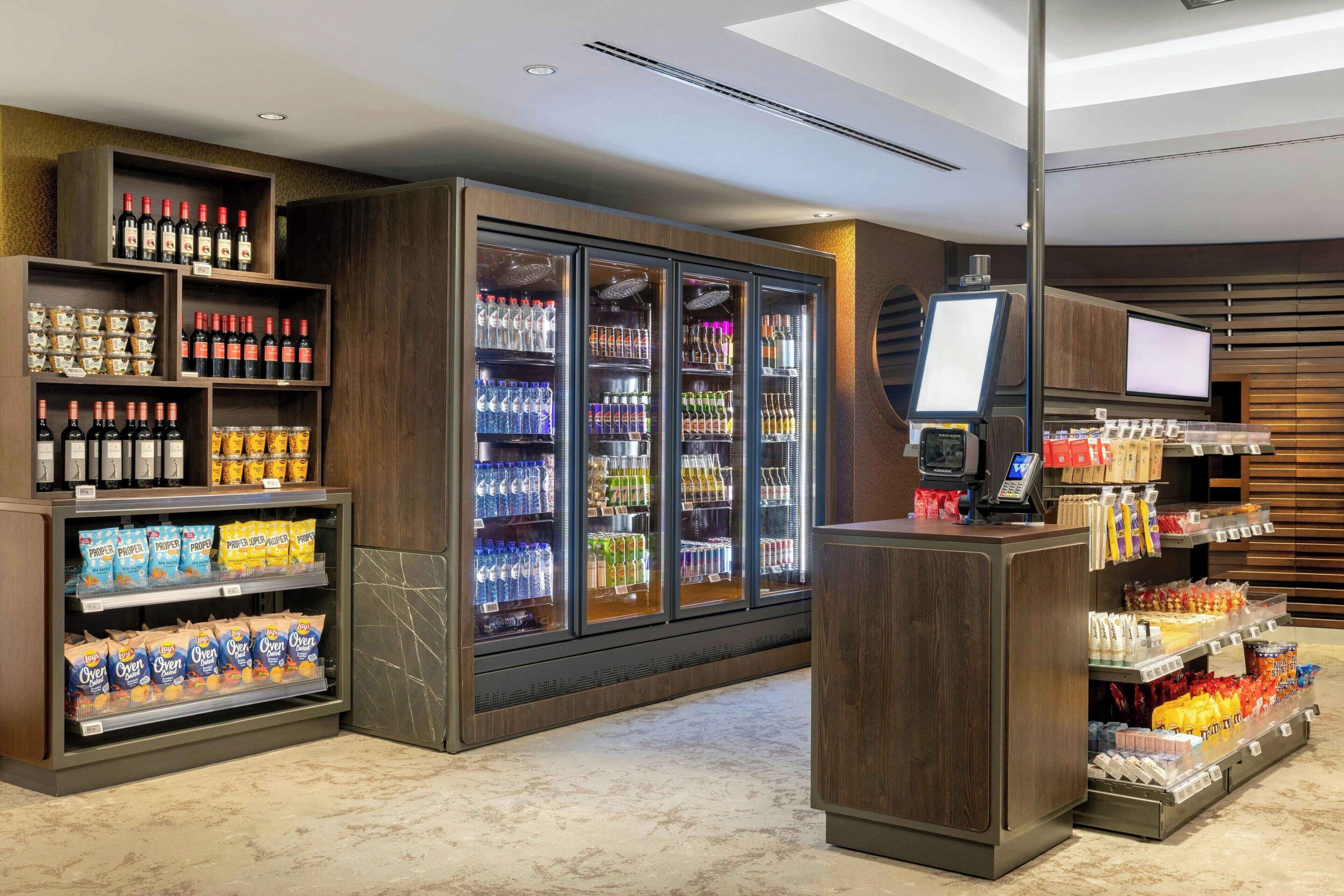 A modern hotel lobby with a self-service beverage station, refrigerated display cases, and a digital screen.