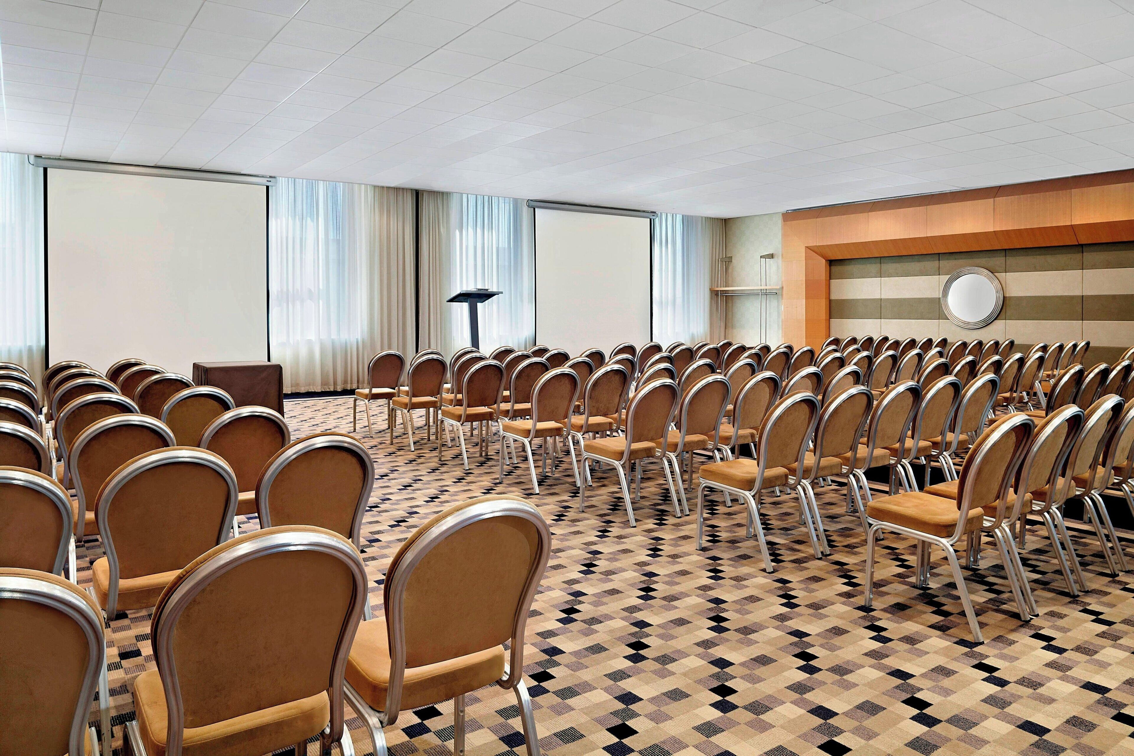 A conference room with rows of chairs, a projector screen, and a large window with curtains.