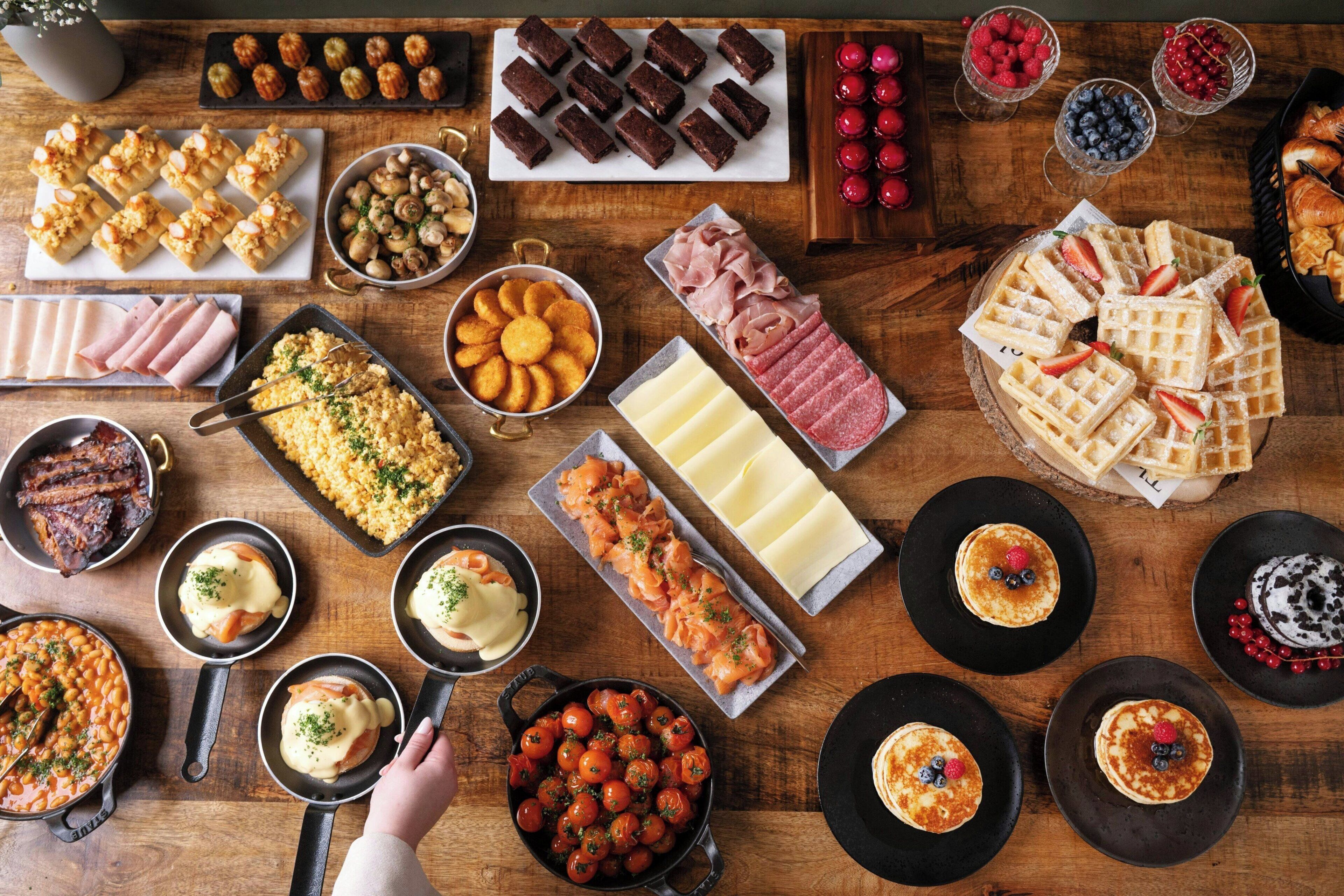 Variedad de alimentos sobre una mesa de madera, incluyendo pasteles, carnes, quesos y frutas.