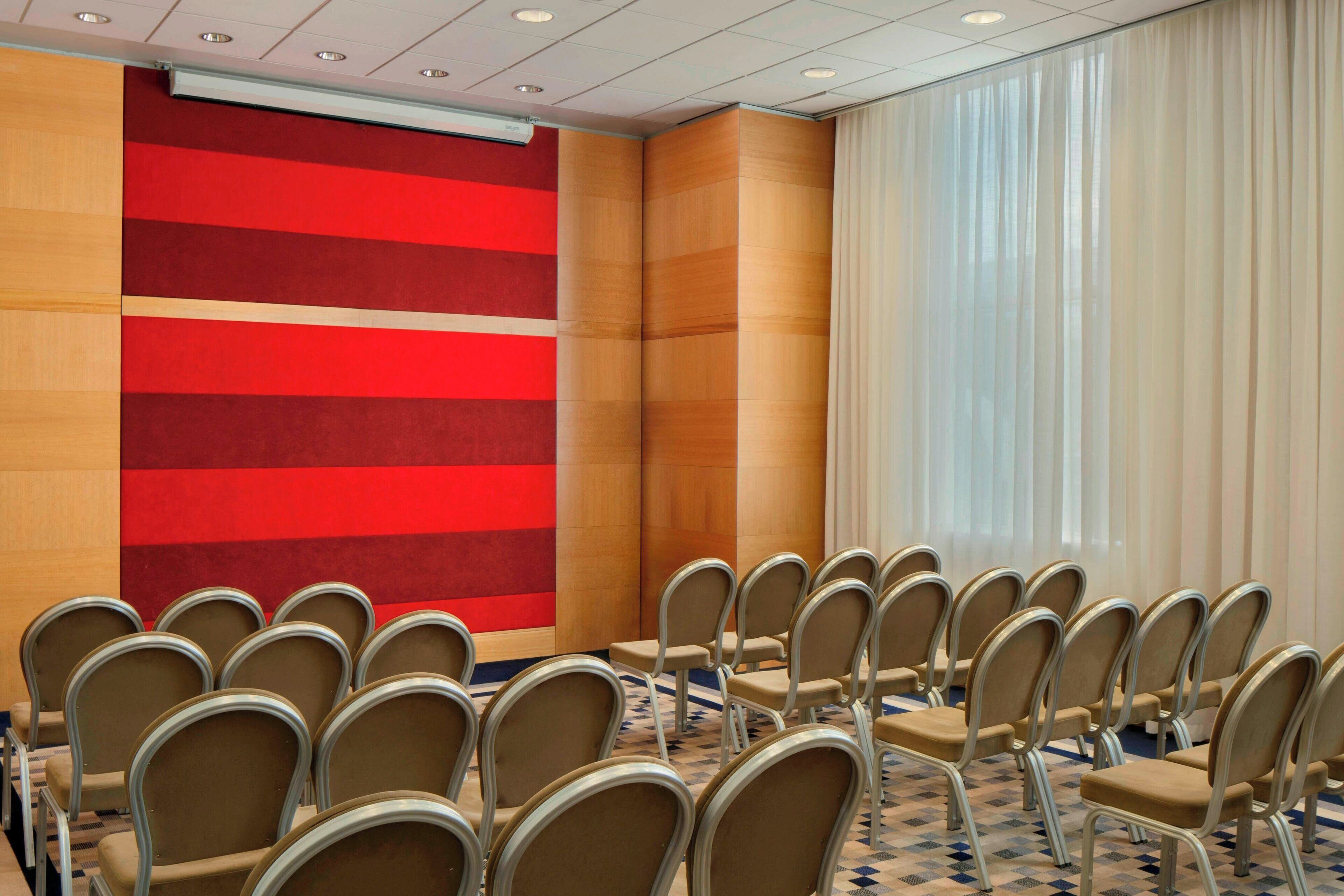 A conference room with rows of beige chairs, a red and beige wall, and a large window with sheer curtains.