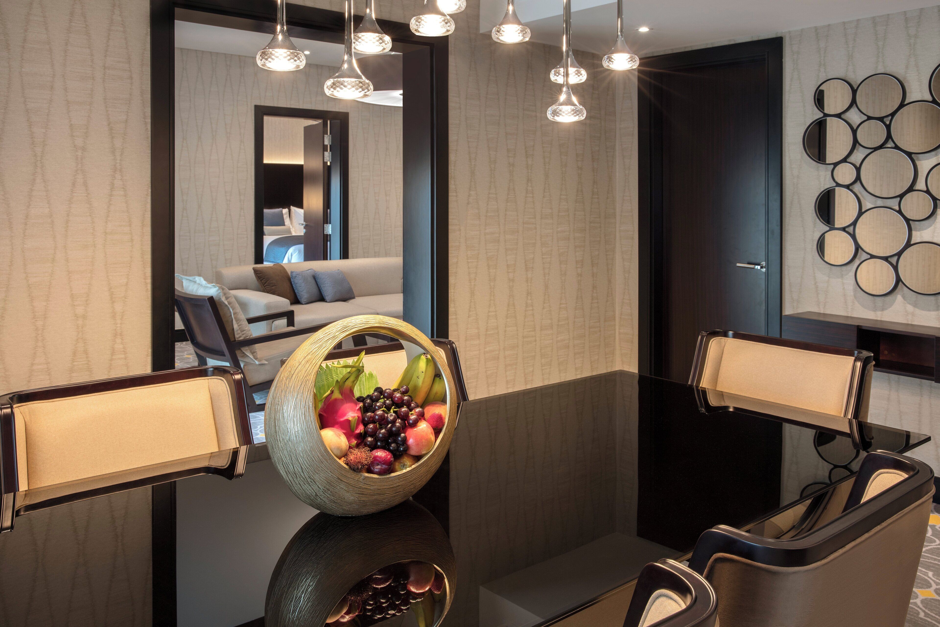 A modern hotel room with a dining table, chairs, a fruit bowl, and a view of the living area.