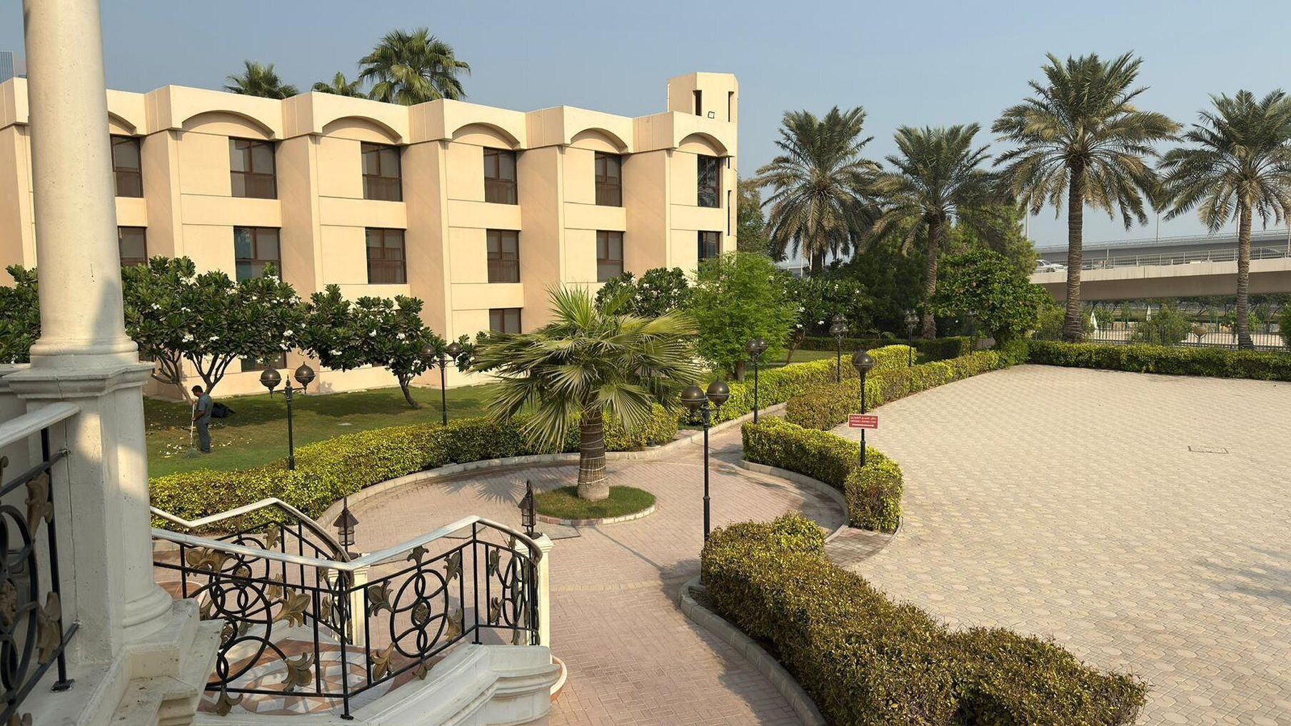 A building with a beige facade, a central courtyard with palm trees, and a paved walkway.