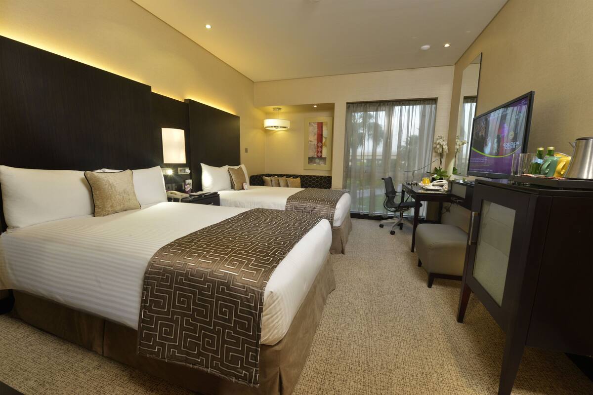 A hotel room with two beds, a desk, a chair, a television, and a bathroom visible through a glass door.
