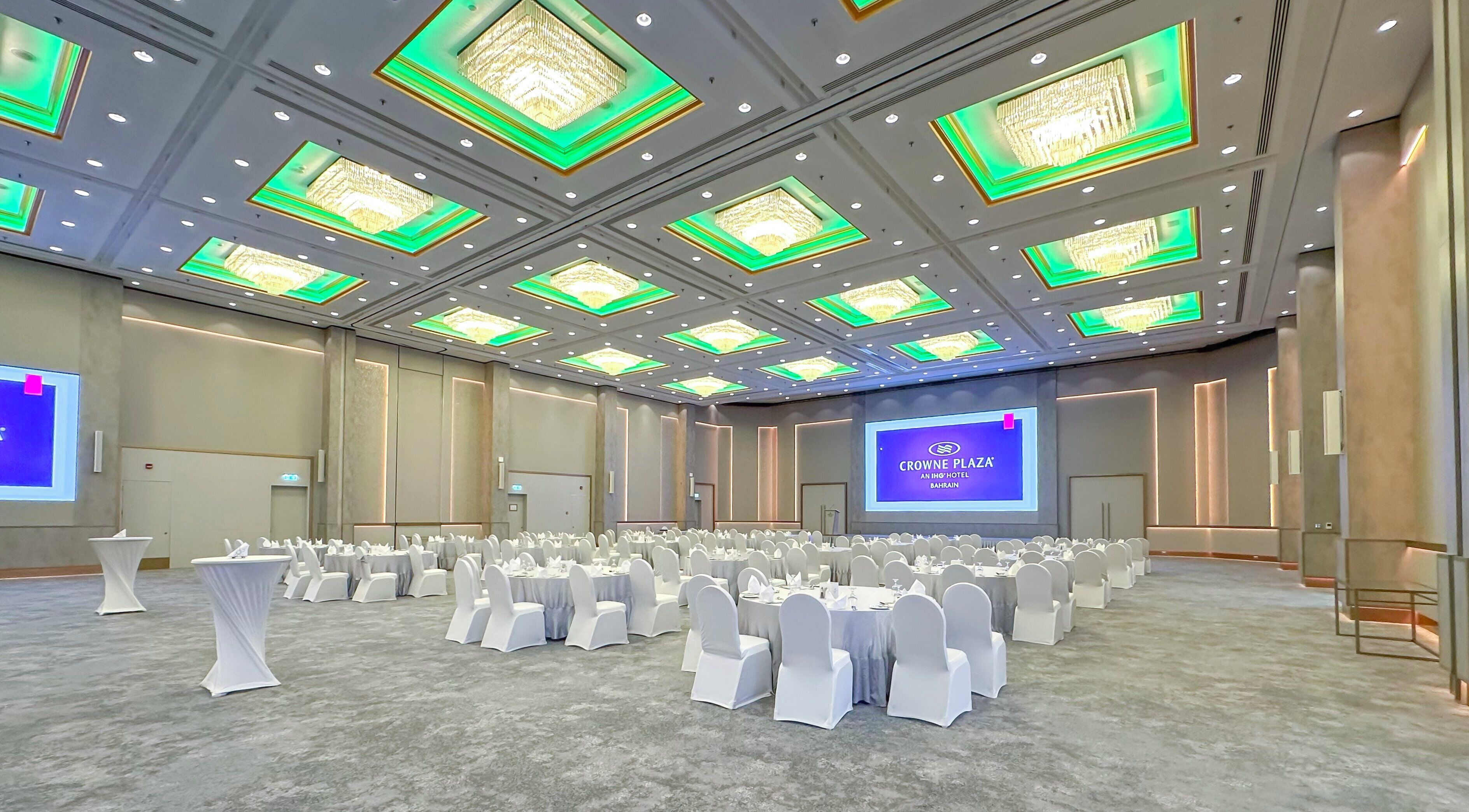 A large conference room with rows of round tables and white chairs, a large screen displaying a presentation, and a chandelier with green lighting accents on the ceiling.