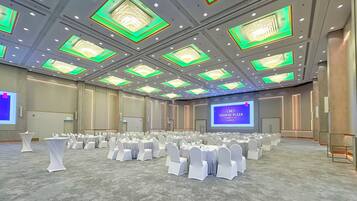 A large conference room with rows of round tables and white chairs, a large screen displaying a presentation, and a chandelier with green lighting accents on the ceiling.