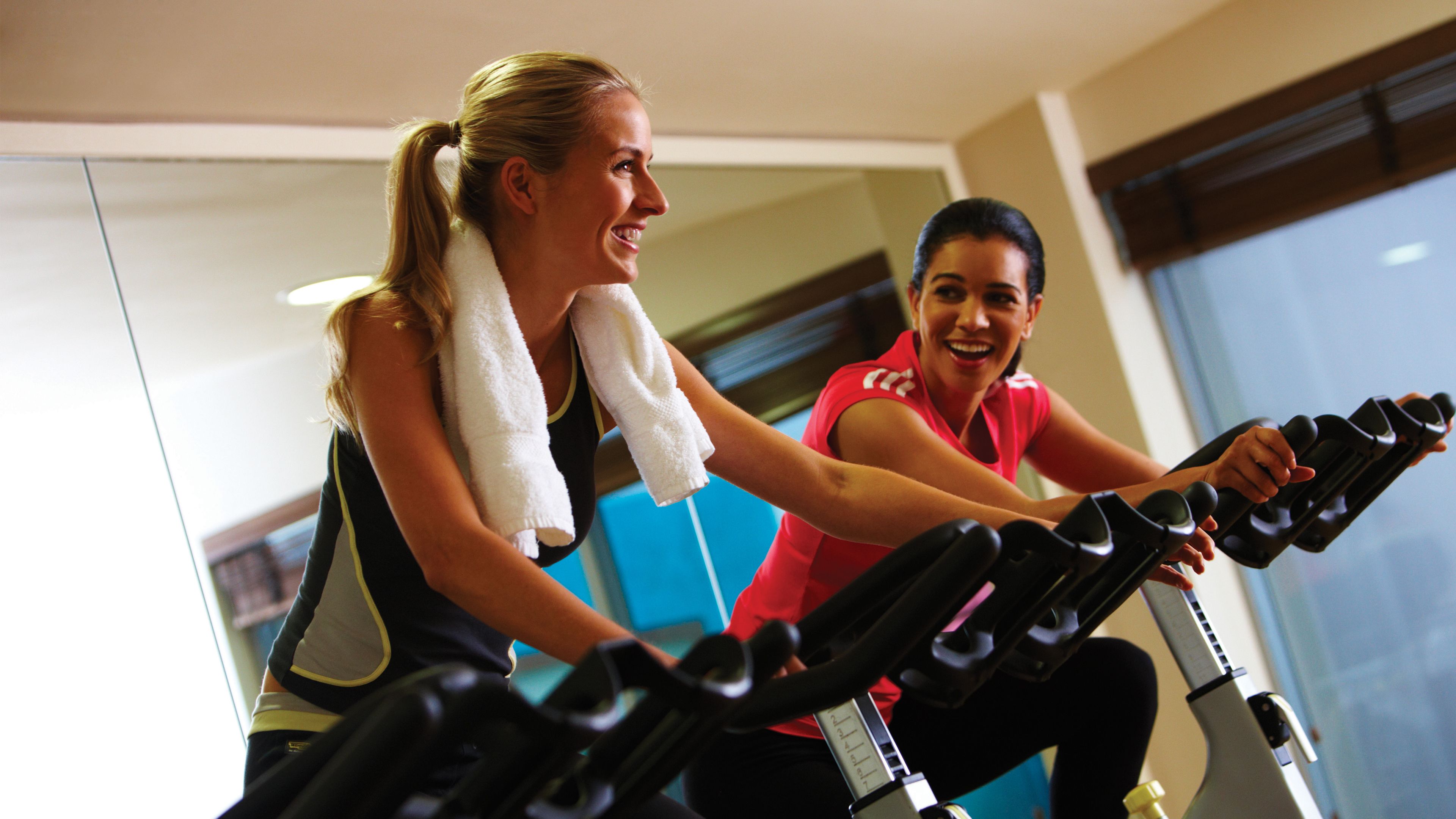 Duas pessoas usando bicicletas ergométricas numa academia com uma janela grande e uma parede azul.