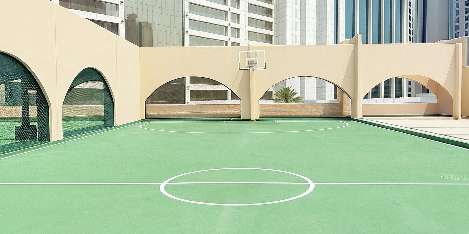 Un terrain de basket en plein air doté d’une surface verte, entouré de murs de couleur beige et agrémenté d’ouvertures en arc de cercle.