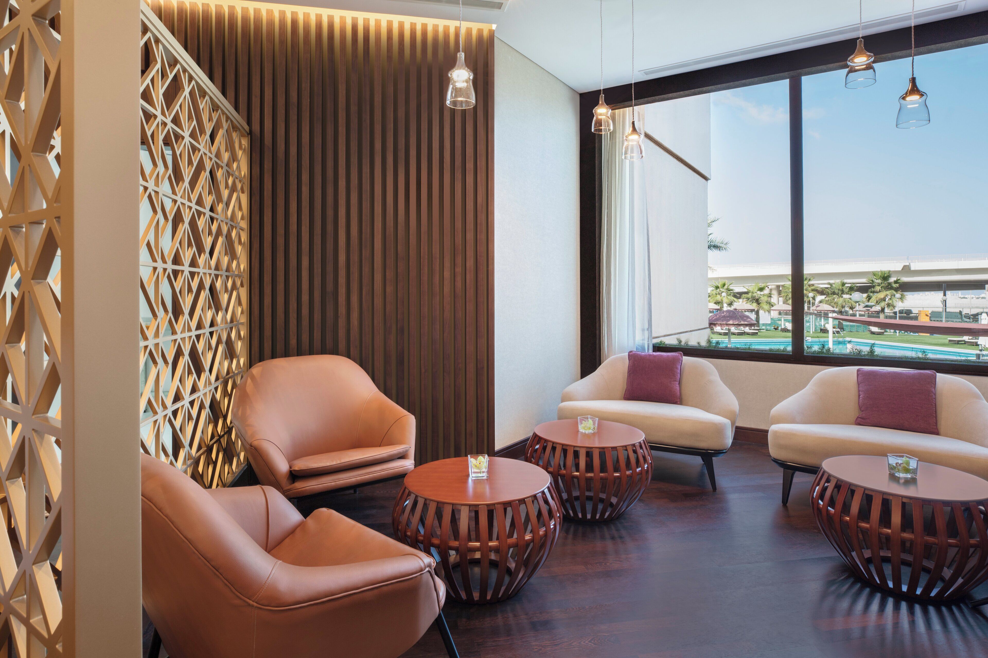 Un moderno espacio de sala de estar con sillones de cuero, mesas redondas de madera y un amplio ventanal con vista a una piscina y palmeras.