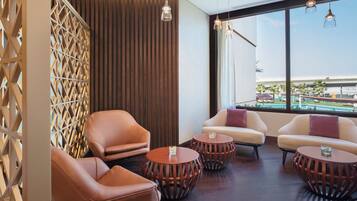 Un moderno espacio de sala de estar con sillones de cuero, mesas redondas de madera y un amplio ventanal con vista a una piscina y palmeras.
