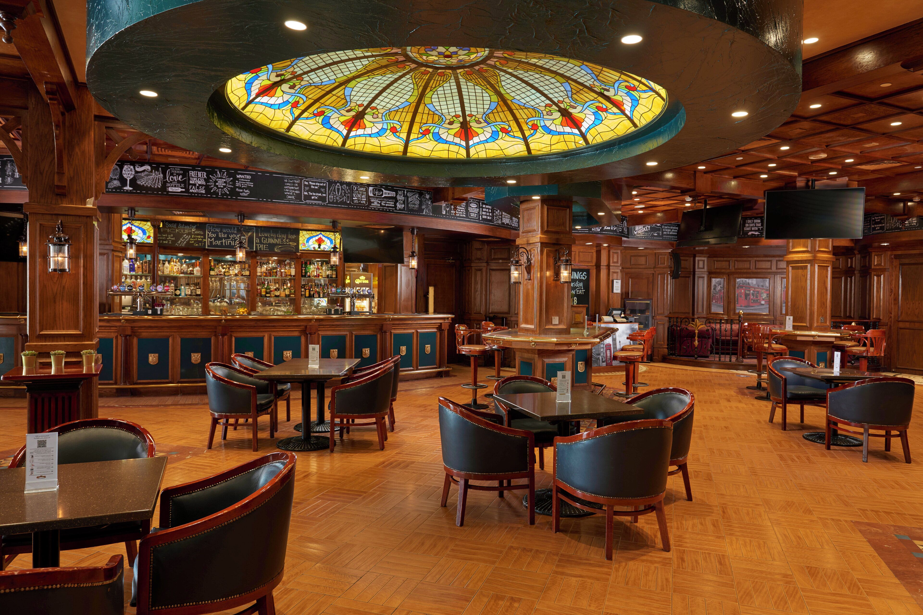A bar area with wooden furniture, a stained glass ceiling, and a bar counter with various bottles.
