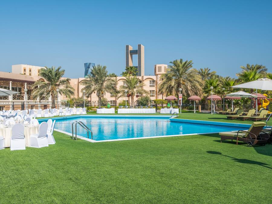 A pool area with a clear blue pool, surrounded by white chairs and tables, palm trees, and a modern building in the background.