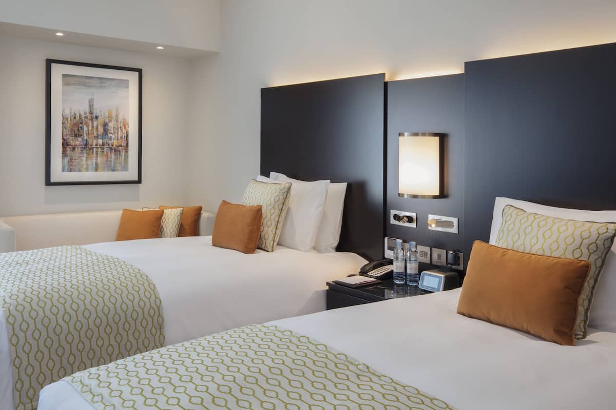 A hotel room with two beds, a cityscape painting, a modern headboard, bedside lamps, and a nightstand with a phone and water bottles.