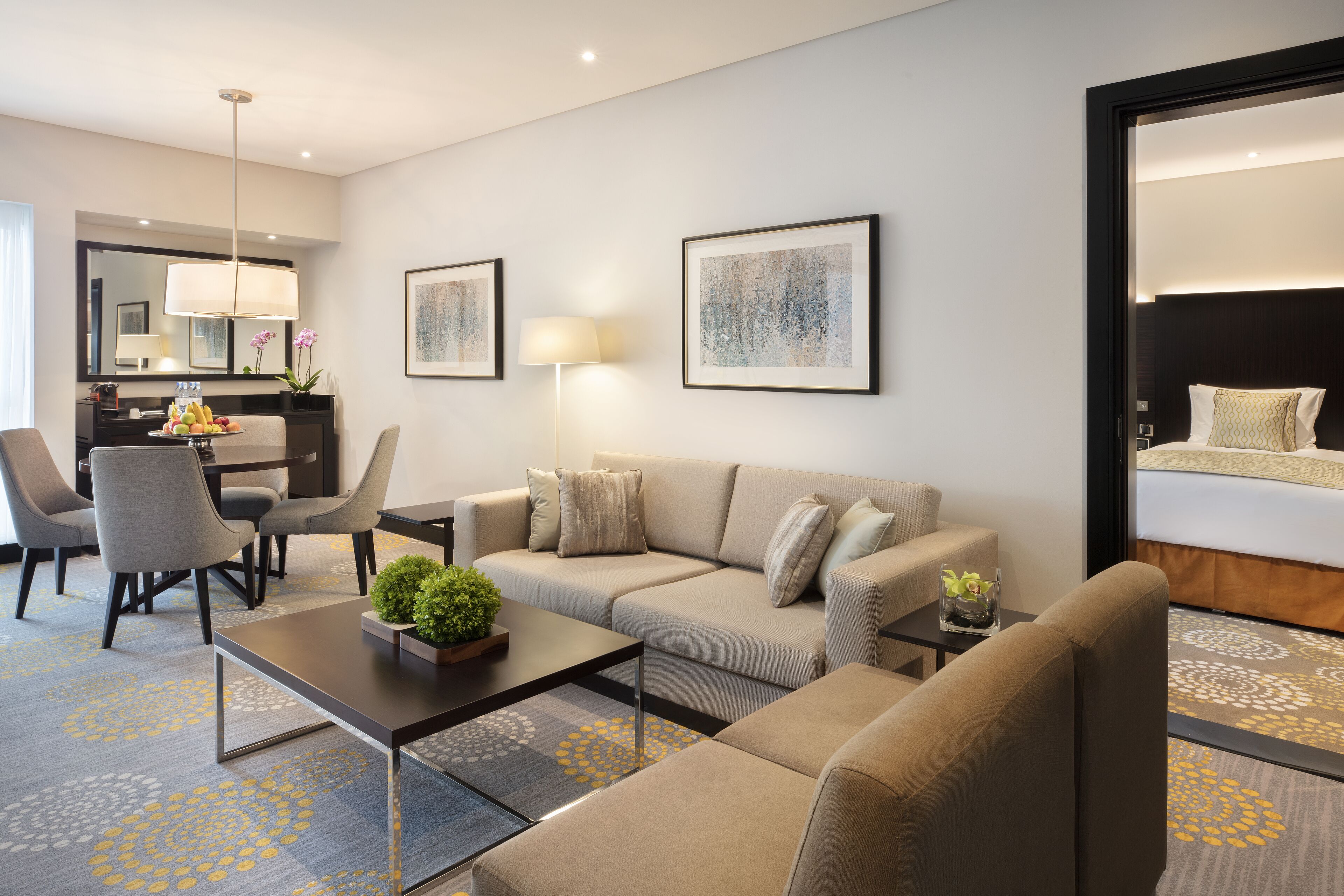 A modern hotel room with a sofa, armchairs, a coffee table, and a dining area with chairs and a lamp.