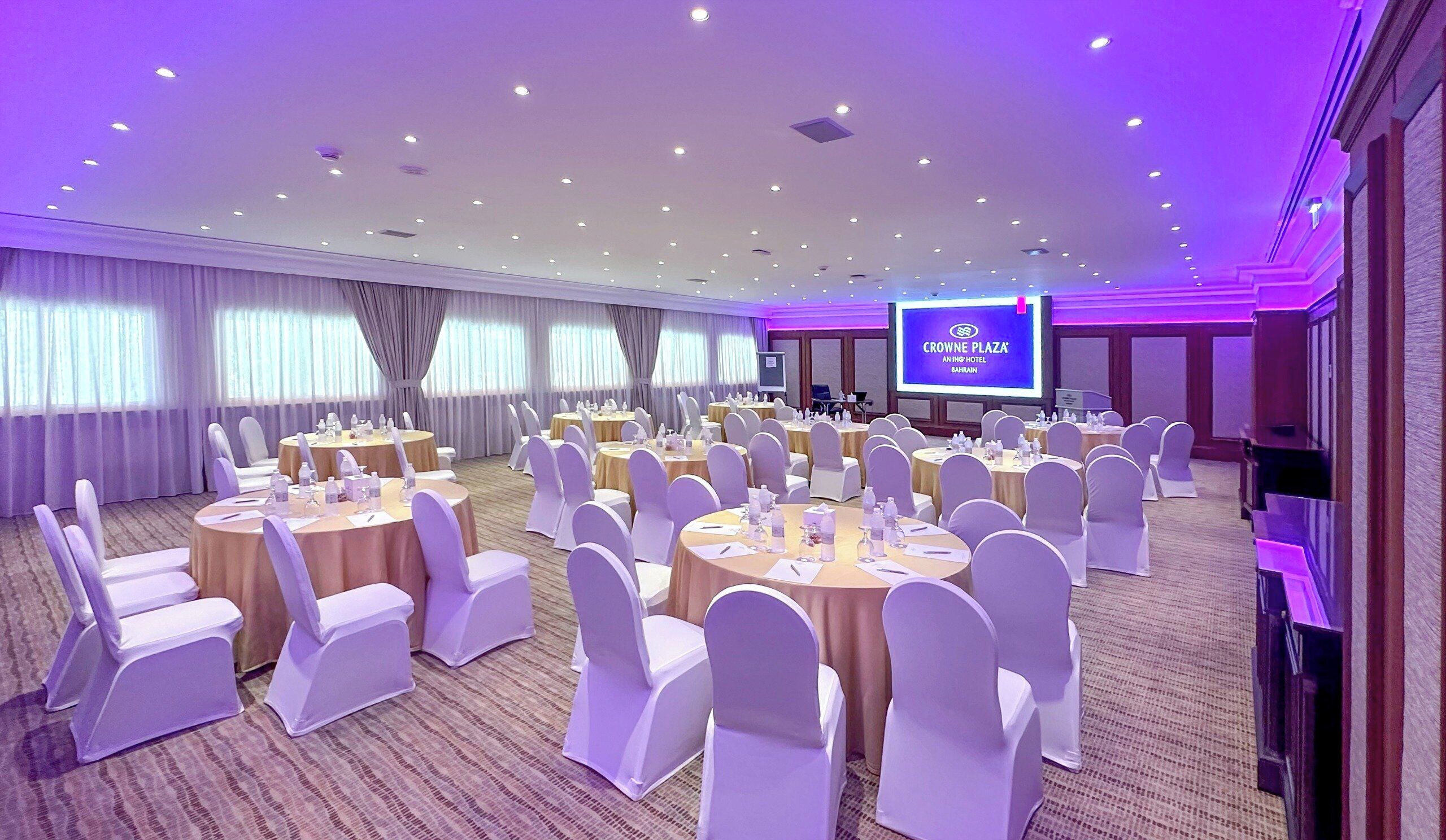A conference room at Crowne Plaza with round tables, white chairs, and a large screen displaying the hotel's name.