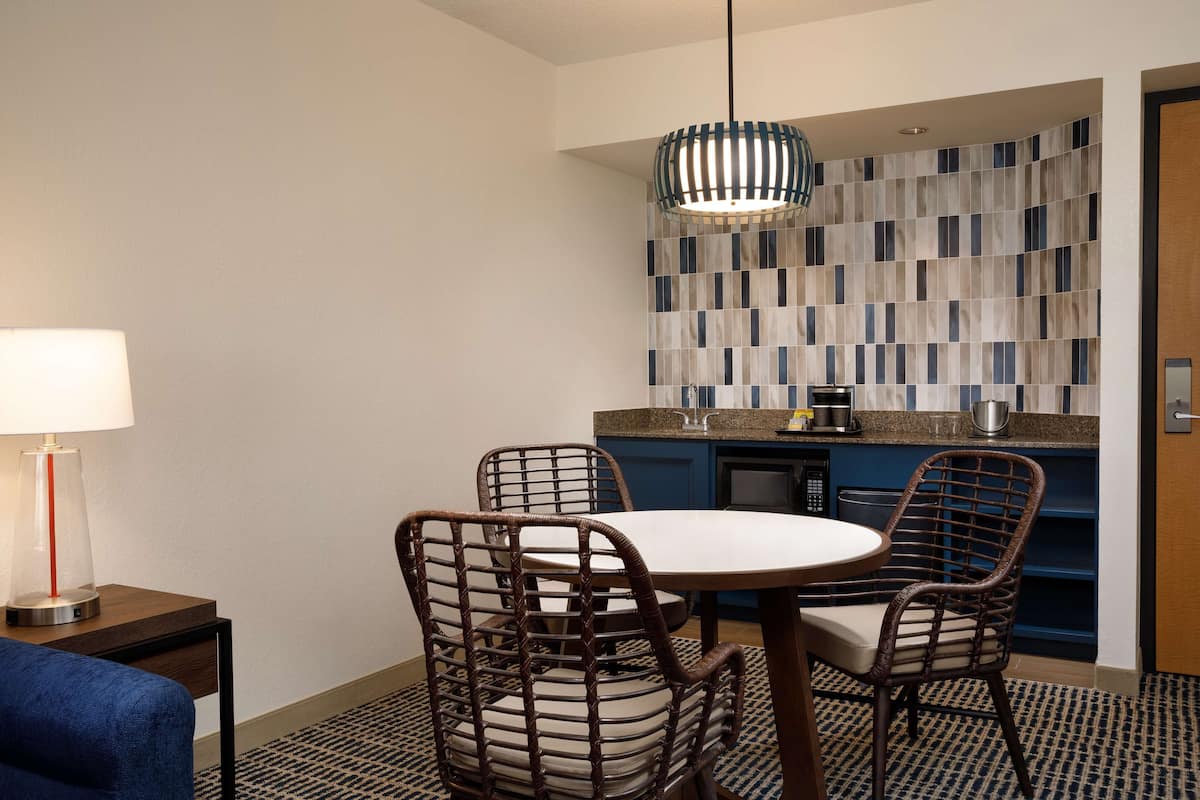 Ein kompaktes Hotelzimmer mit Esstisch, zwei Korbstühlen, einer Lampe, einer Küchenzeile mit blauer Spritzschutz und einem gemusterten Teppich.