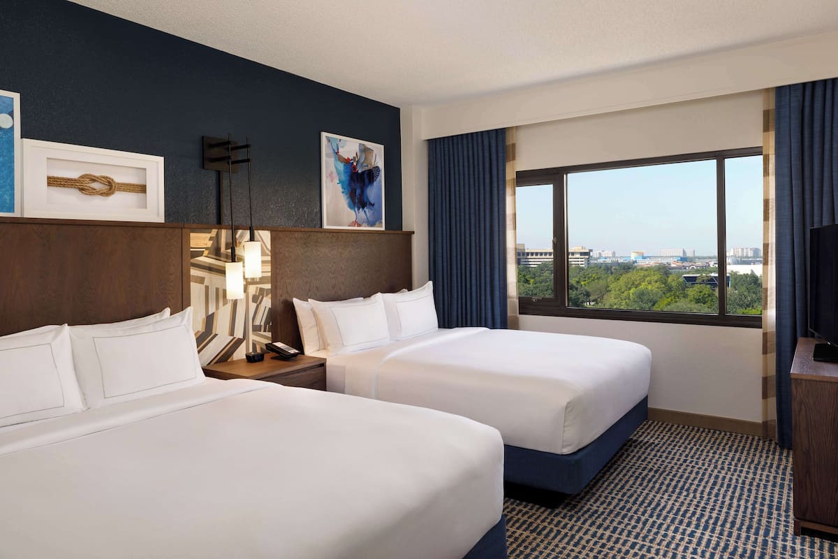 Ein Hotelzimmer mit zwei Betten, einem großen Fenster mit Blick auf die Stadt, einem Flachbildfernseher und einem Schreibtisch mit Lampe.