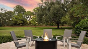 A circular outdoor fire pit surrounded by four chairs, set on a paved patio with a view of a golf course and trees in the background.