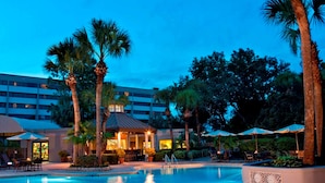A swimming pool with lounge chairs and umbrellas, surrounded by palm trees and a building in the background.