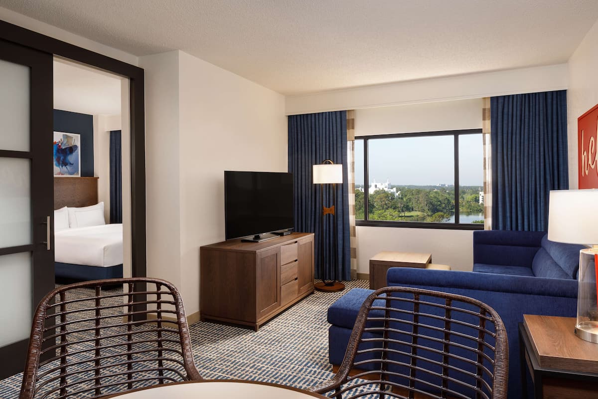 Ein Hotelzimmer mit einem Bett, einem Fernseher, einem Sofa, zwei Korbstühlen, einem kleinen Tisch und einem Fenster mit Blick auf Grünflächen.