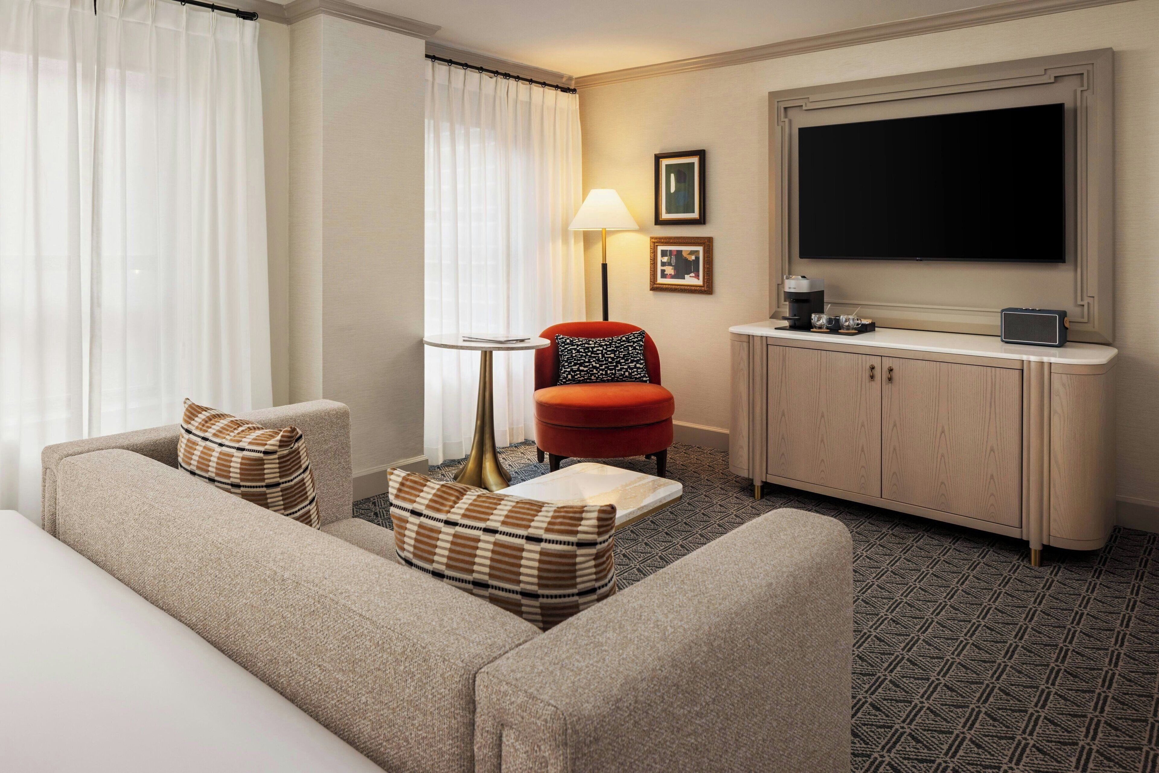 A hotel room with a sofa, armchair, ottoman, lamp, TV, and a cabinet.