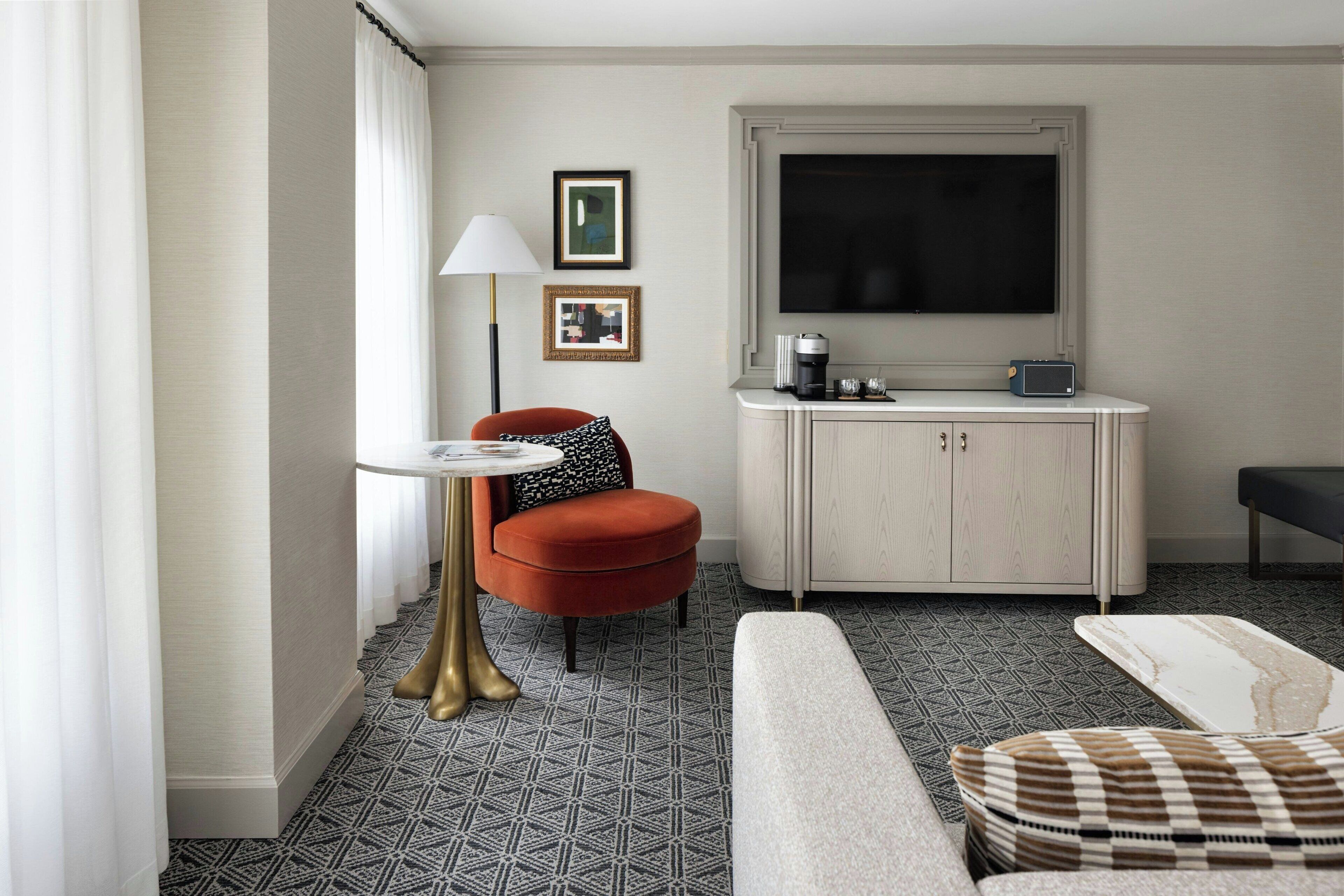 A modern hotel room with a sofa, a small round table, a red armchair, a cabinet with a flat-screen TV, and a patterned floor.