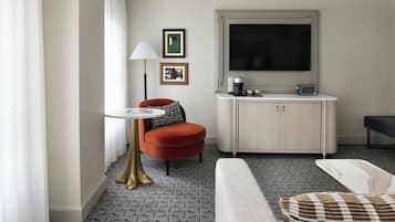 A modern hotel room with a sofa, a small round table, a red armchair, a cabinet with a flat-screen TV, and a patterned floor.