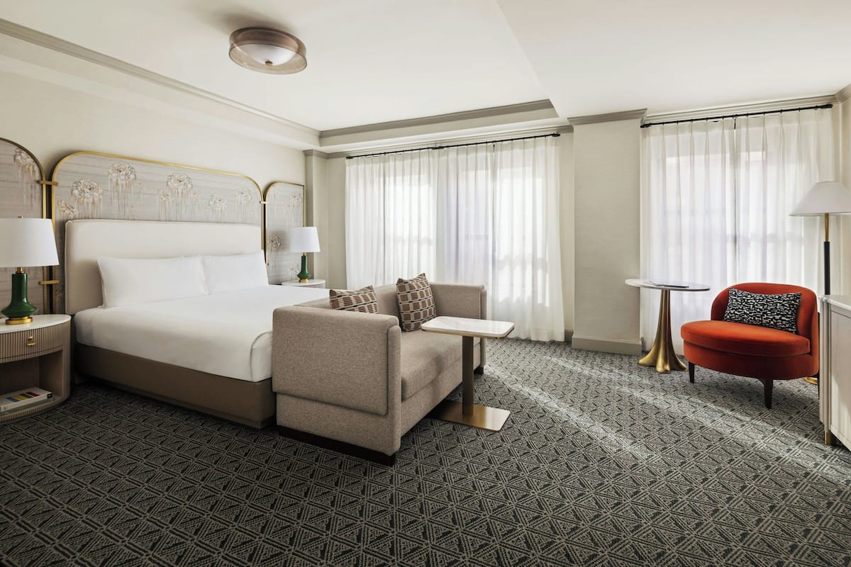 A hotel room with a bed, a sofa, a small table, a chair, and a lamp.