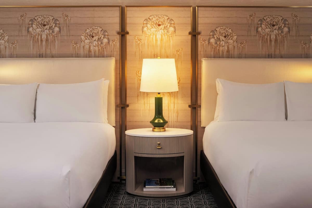 A hotel room with two beds, a round nightstand with a lamp, and a bookshelf.