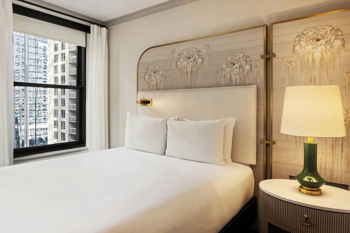 A hotel room with a large bed, a nightstand, a lamp, and a view of a cityscape through a window.