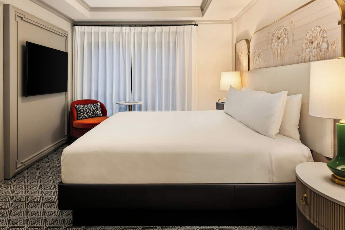 A hotel room with a large bed, a flat-screen TV mounted on the wall, a red chair, a small round table, and a lamp on a bedside table.