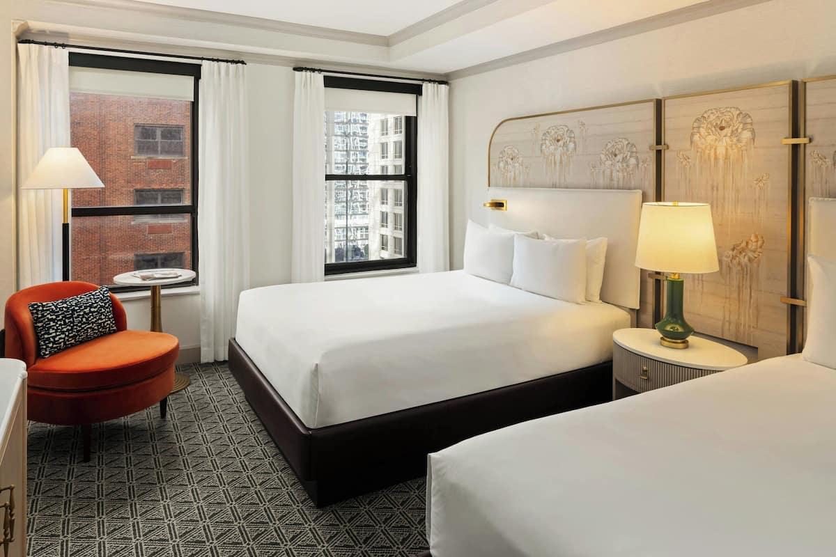 A hotel room with two beds, a red armchair, a small round table, and a view of a brick building through the window.