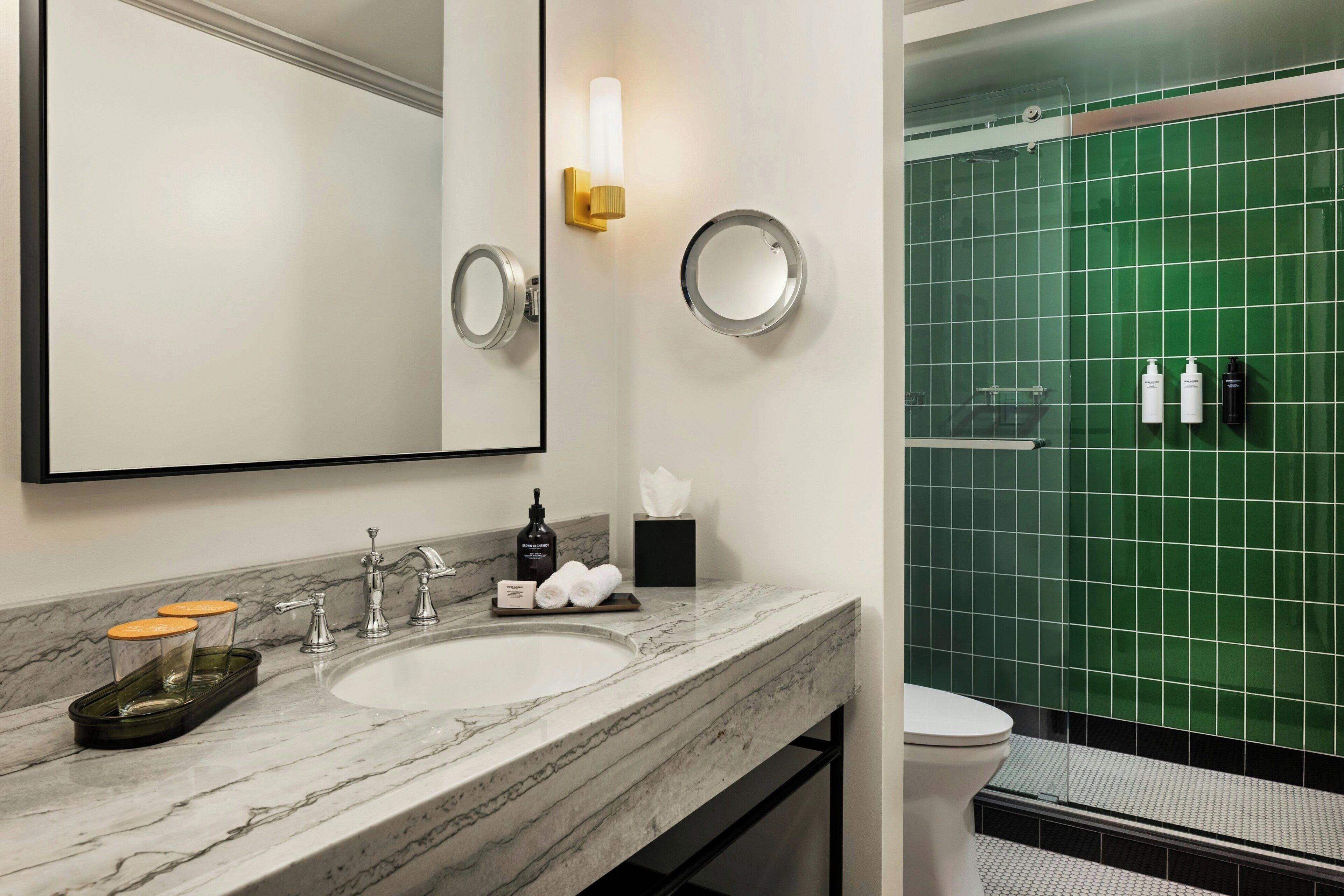 Une salle de bain avec un lavabo en marbre, deux miroirs, un porte-serviettes et une zone de douche carrelée de carreaux verts.