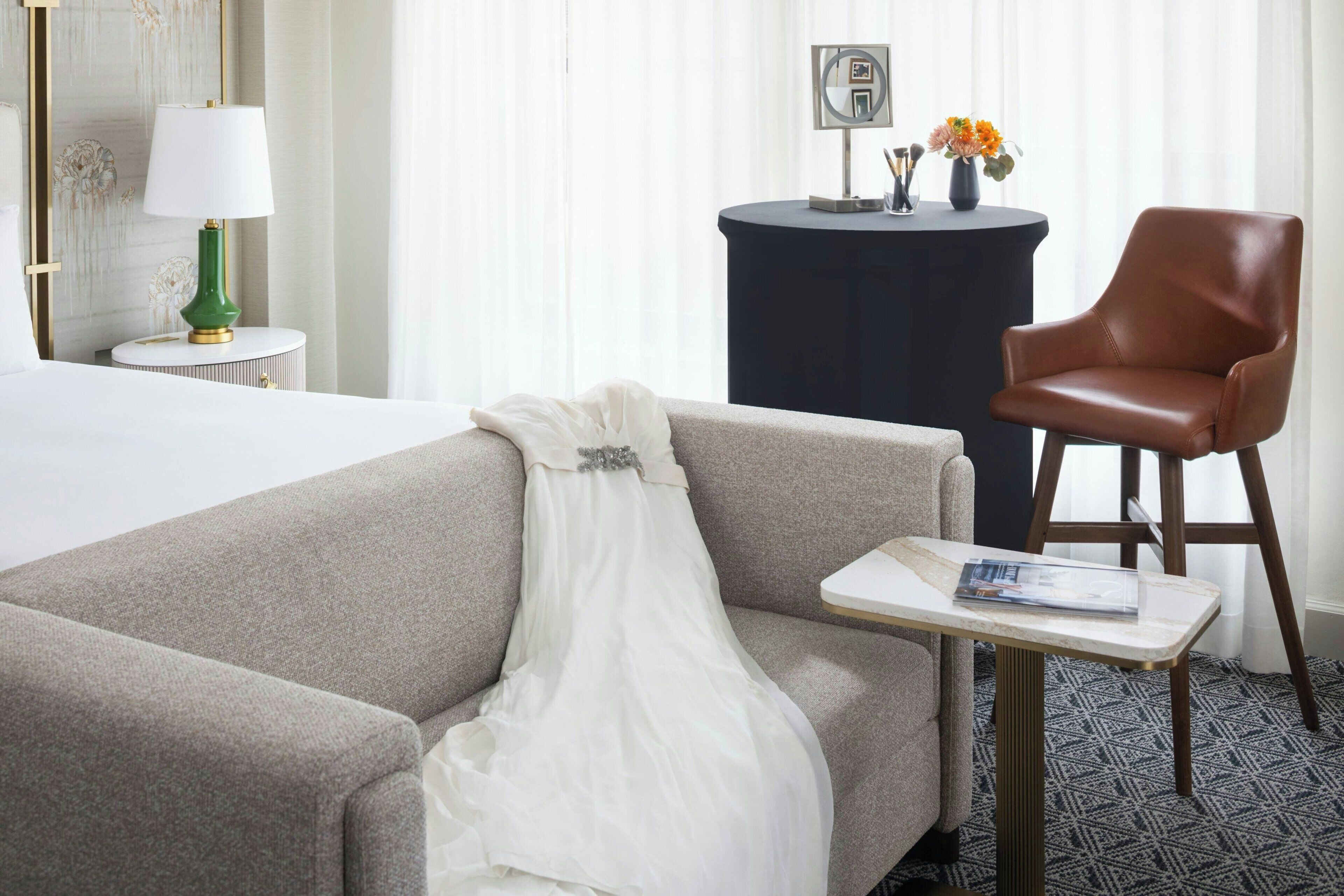 Una habitación de hotel con cama, sofá, una mesita con una revista, lámpara, sillón y una mesa redonda con un florero.