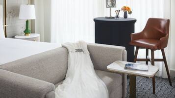 Una habitación de hotel con cama, sofá, una mesita con una revista, lámpara, sillón y una mesa redonda con un florero.
