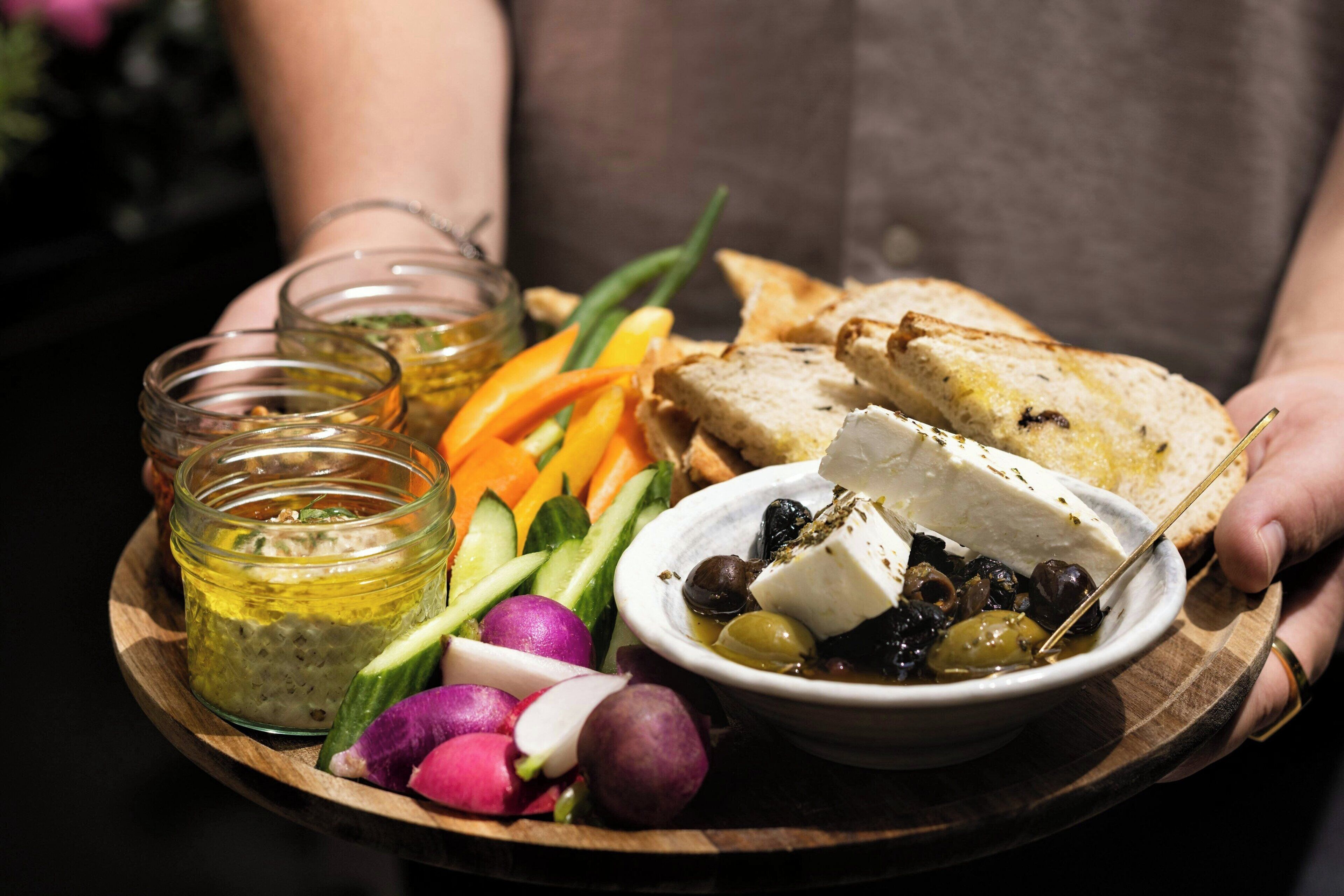 A wooden board with a variety of foods including cheese, olives, radishes, and bread.