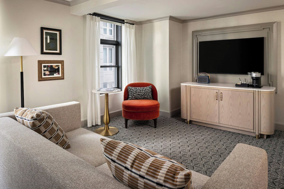 A hotel room with a sofa, a red chair, a small table, a TV, and a cabinet.