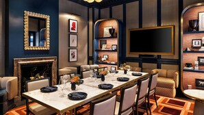A dining area with a long table, chairs, and a fireplace. There are framed pictures on the walls and a large flat-screen TV mounted on the wall.