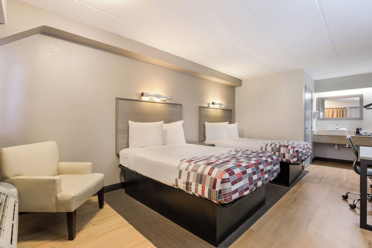 Ein Hotelzimmer mit zwei Betten, einem Sessel, einem Schreibtisch und einem Spiegel.