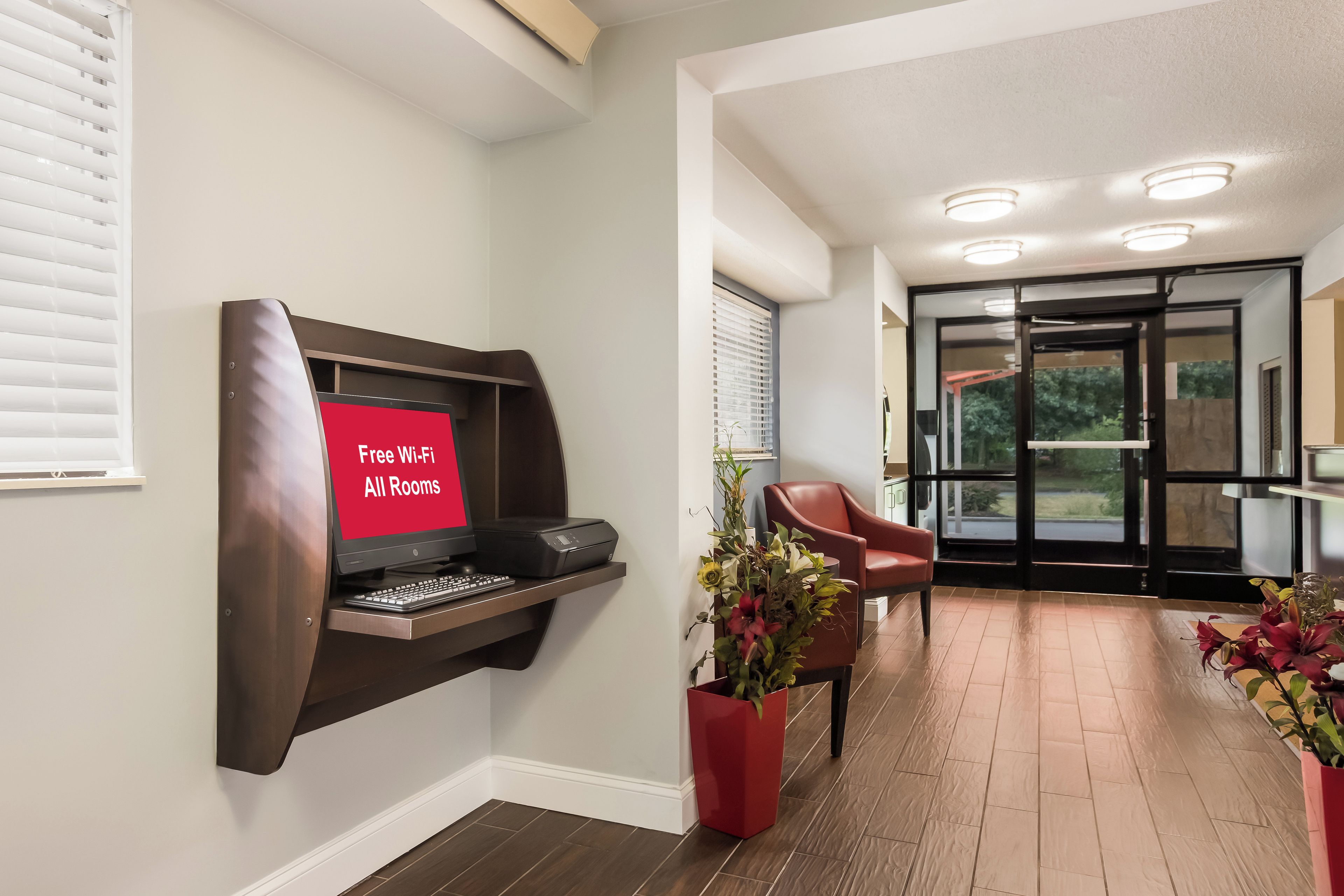 A hotel room with a desk, a printer, a red sign indicating free Wi-Fi, a red chair, and a potted plant.