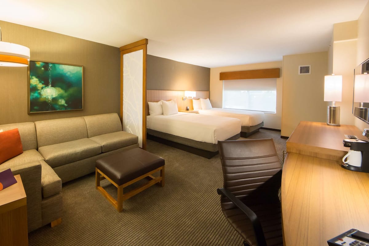 A hotel room with a sofa, a bed, a desk, a chair, a coffee machine, and a painting on the wall.