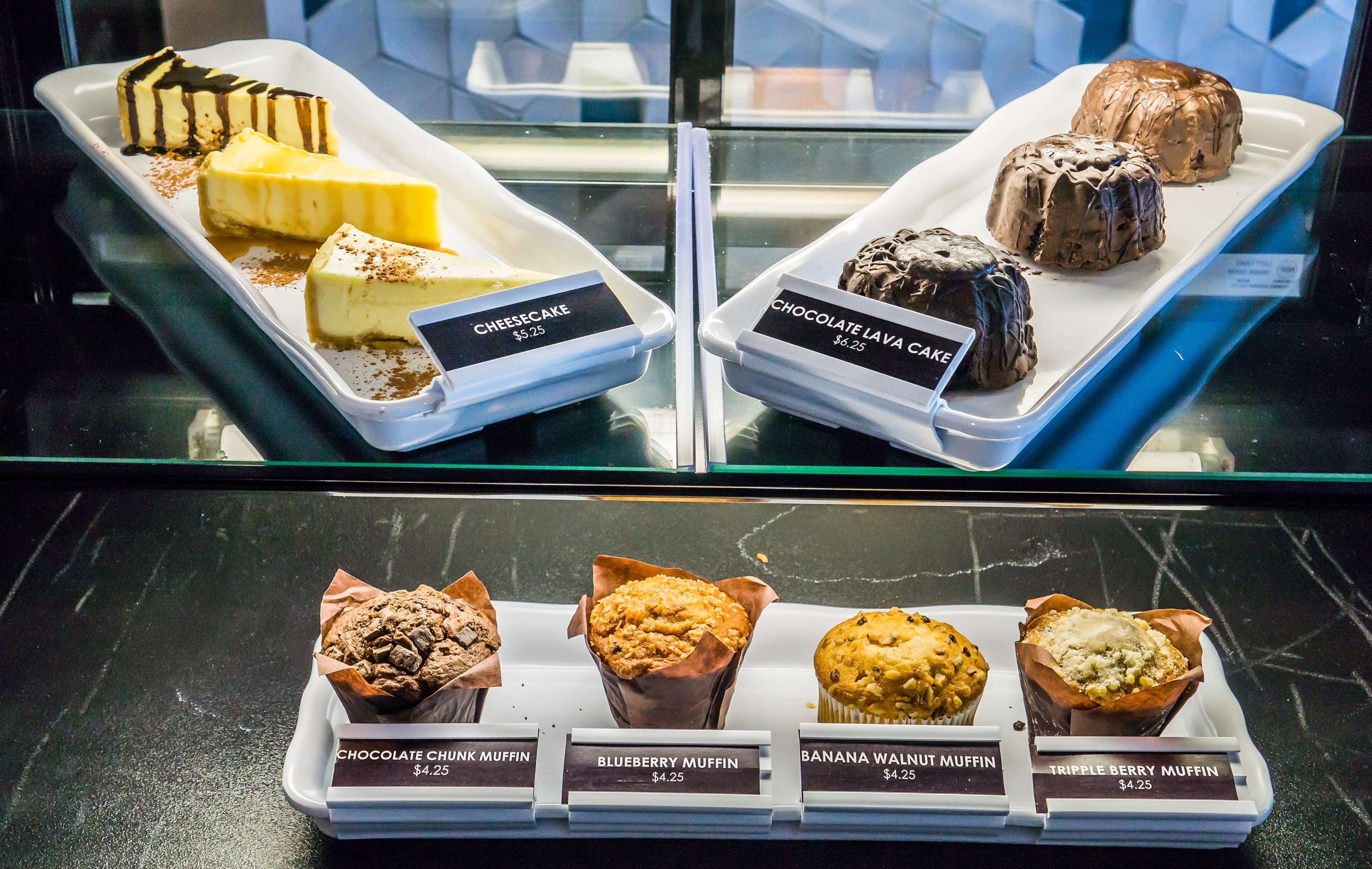 A display case with various desserts and muffins, including cheesecake, chocolate lava cake, chocolate chunk muffin, blueberry muffin, banana walnut muffin, and triple berry muffin, each priced at $4.25.