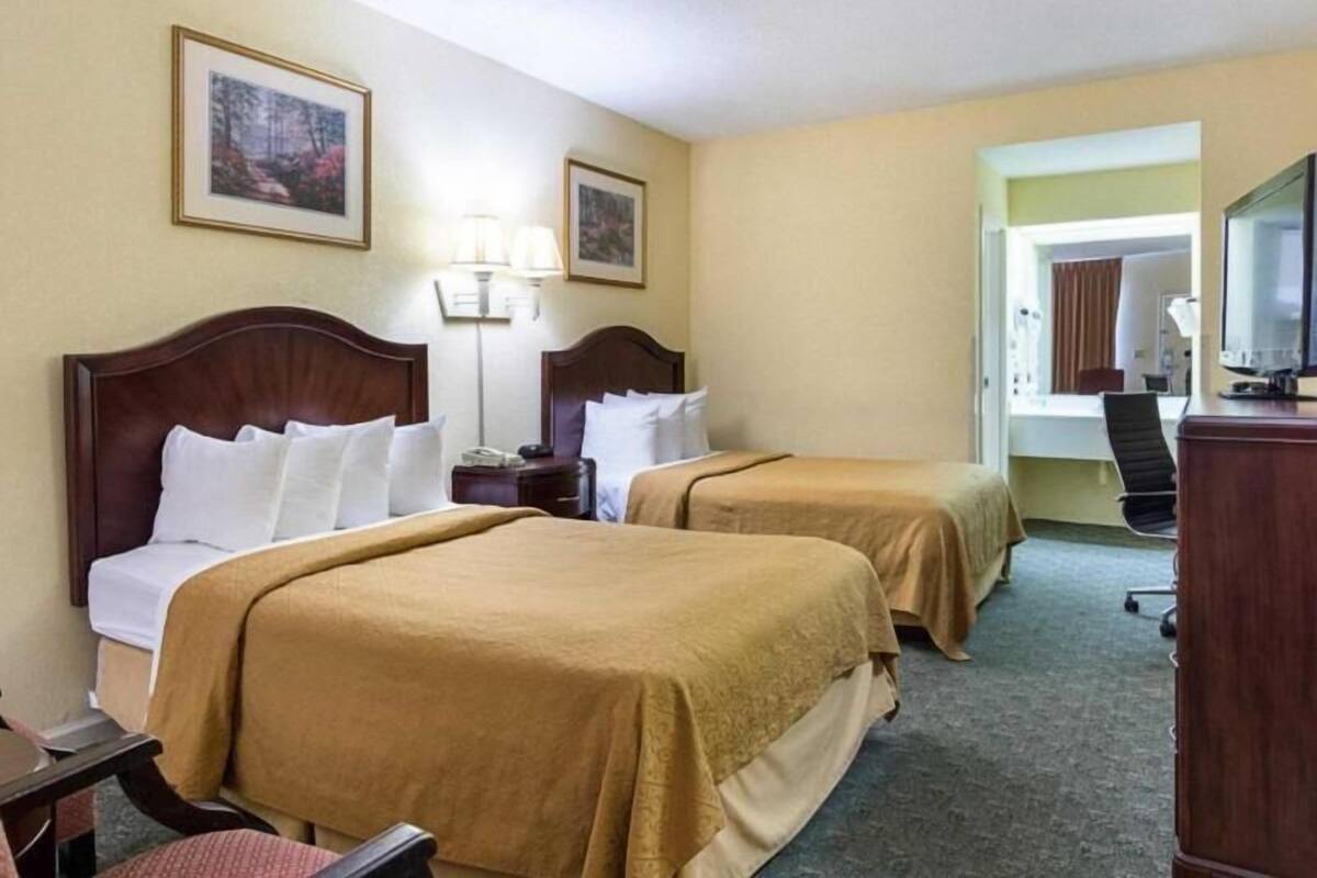 A hotel room with two beds, a desk, a chair, a lamp, and a TV.