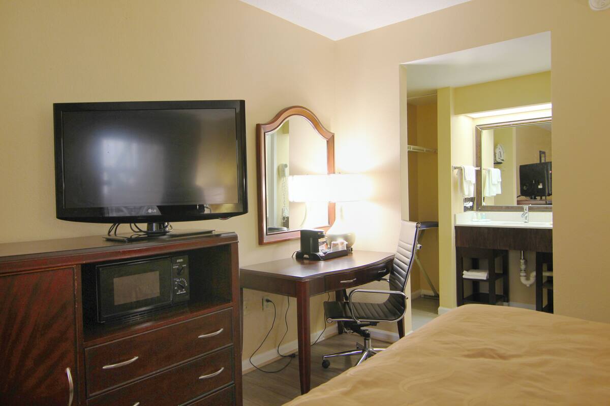 A hotel room with a flat-screen TV, a desk with a lamp, a chair, a bed, and a bathroom with a mirror and sink.